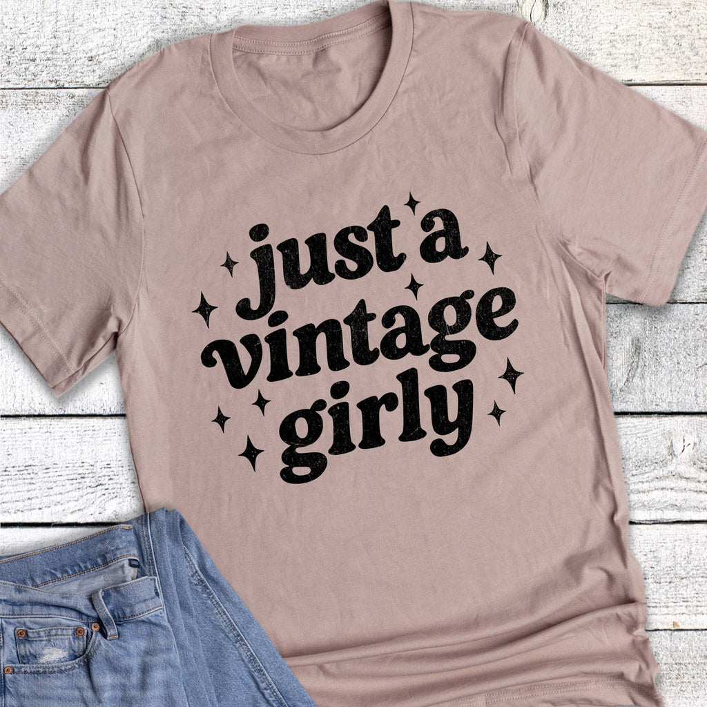 Light-colored graphic tee with 'just a vintage girly' text, featuring a relaxed fit, paired with denim shorts.