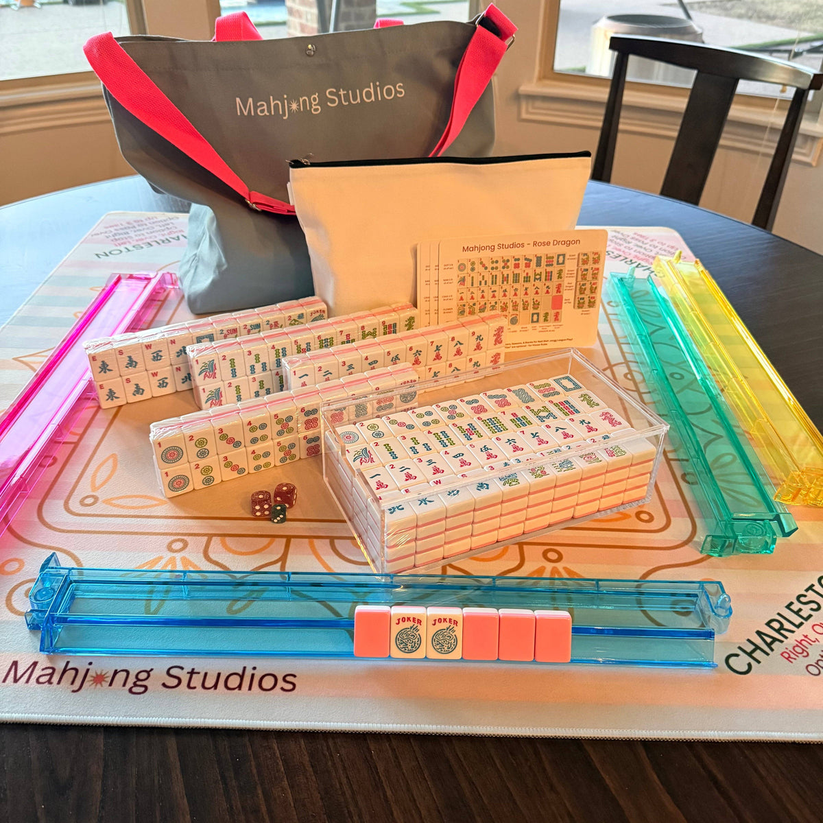 A colorful Mahjong tile set featuring pink and clear racks, dice, and a stylish carrying bag, arranged on a printed mat.