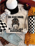 Light gray sweatshirt featuring the text 'THE BAGS UNDER MY EYES ARE DESIGNER' with a stylish graphic design.