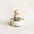 Round speckled stoneware container with solid dish soap and a wooden brush on top, designed for effective dish cleaning.