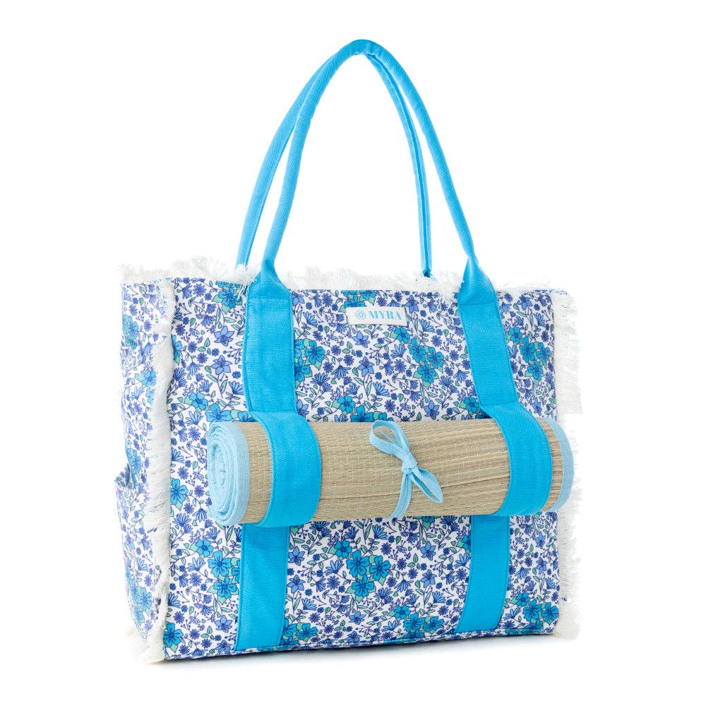 Shoreline Weekender Bag featuring a wildflower print body, fringe detailing, blue carry straps, and a rolled beach mat.