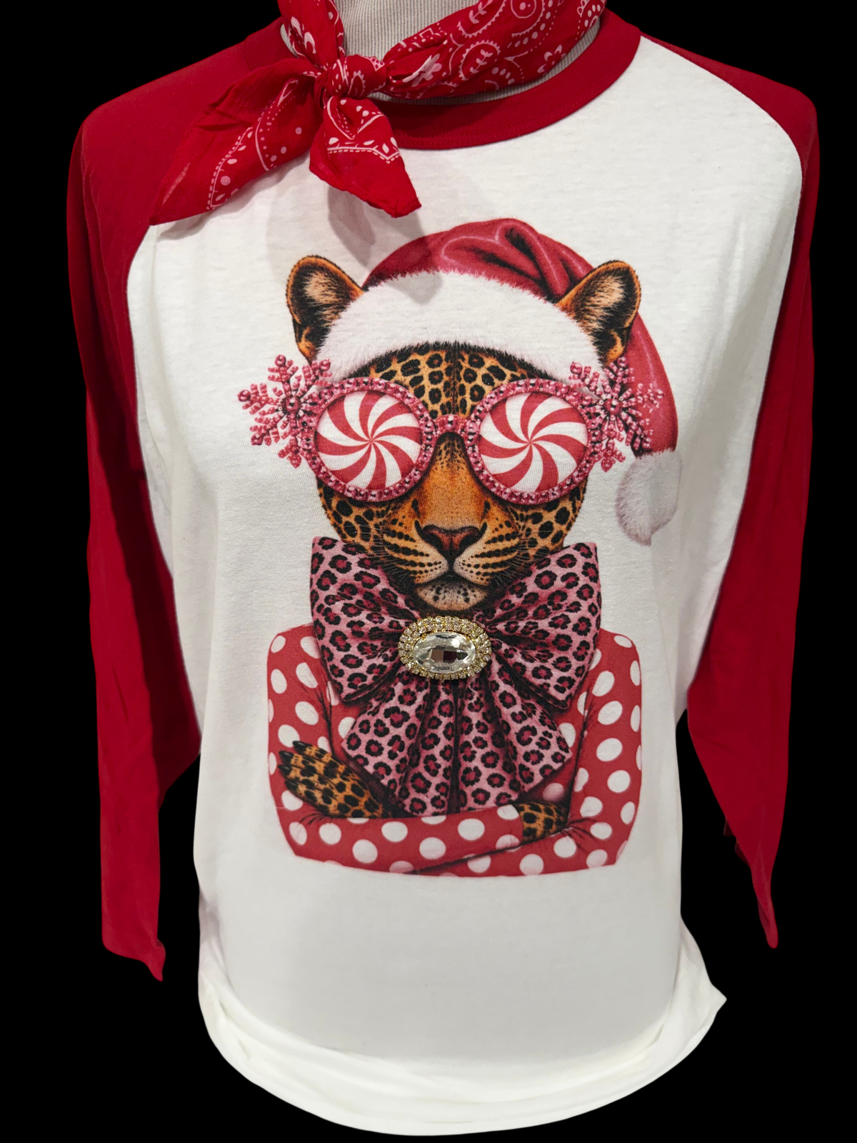 A peppermint leopard wearing a Santa hat and oversized sunglasses on a white and red raglan shirt with a rhinestone brooch.